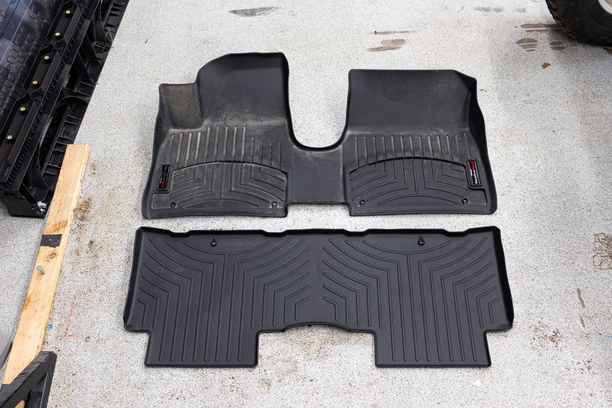 A pair of all-weather rubber floor mats leaning against a driveway curb, freshly rinsed and drying in sunlight